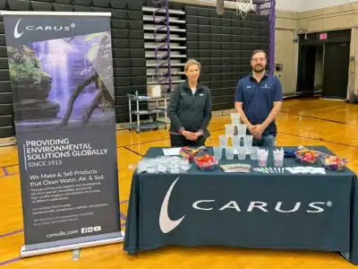 Two people stand behind a table with a "Carus" logo at a gym event. The table has informational materials, small cups, and bowls of candy. Next to them is a tall banner about environmental solutions by Carus since 1915.