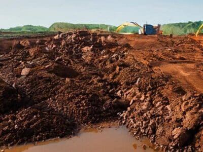 A construction site with large excavation machinery working on a muddy landscape with piles of dirt and a partially dug trench. In the background, green trees and a clear sky are visible.