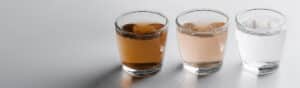 Three glasses of water are lined up on a reflective surface. The first glass contains brown water, the second glass has slightly cloudy water, and the third glass holds clear water.