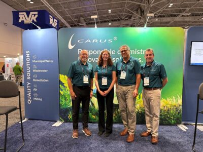 Four people in teal shirts and black pants stand smiling in front of a green and blue Carus company booth display at a convention center. The display lists solutions for water, wastewater, remediation, air, and mining.