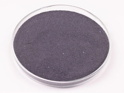 A clear glass petri dish filled with fine, dark gray powder, sitting on a white surface.