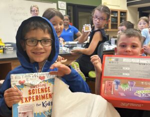 Children participating in Science FUN’damentals, sponsored by Carus & the Illinois Valley Community College’s Continuing Education Department appear excited with new science-themed gifts. A boy in the front smiles, holding up a science experiment book, while another holds a science kit.