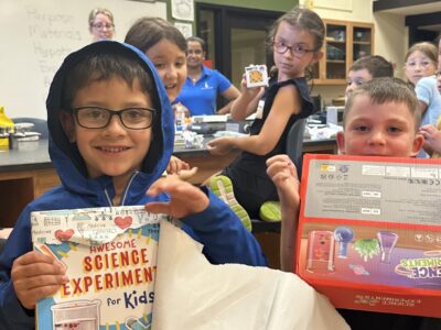 Children participating in Science FUN’damentals, sponsored by Carus & the Illinois Valley Community College’s Continuing Education Department appear excited with new science-themed gifts. A boy in the front smiles, holding up a science experiment book, while another holds a science kit.