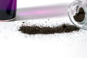 A small glass vial tipped over, spilling a pile of fine, dark powder onto a white surface. In the background, there is a container with a purple liquid, slightly out of focus.