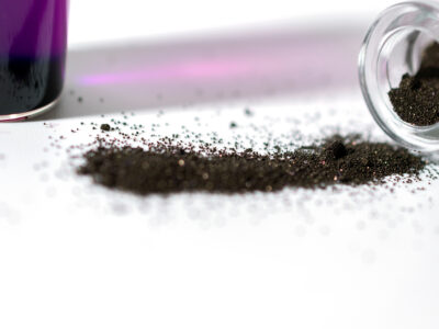 A small glass vial tipped over, spilling a pile of fine, dark powder onto a white surface. In the background, there is a container with a purple liquid, slightly out of focus.