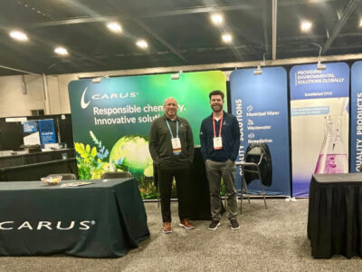 Two men stand at a trade show booth for Carus, featuring banners that promote responsible chemical and innovative solutions. The booth displays information, a bowl of candy, and branded materials on tables.