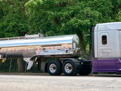 A shiny silver tanker truck with a purple and gray cab is parked on a paved street beside a fence and lush green trees on a sunny day.
