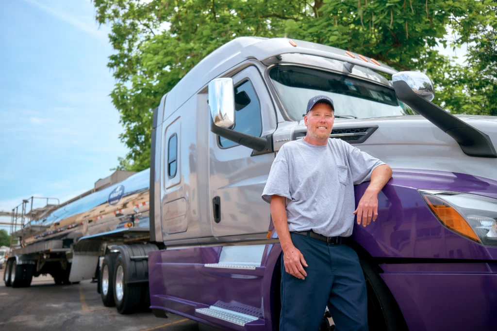 A man in a gray T-shirt and navy pants stands smiling beside a large silver and purple semi-truck and tanker trailer, with green trees and blue sky in the background.