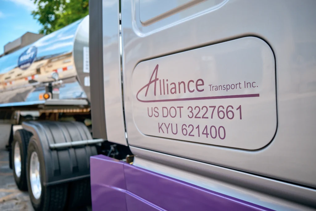 Close-up of a truck door displaying "Alliance Transport Inc." along with US DOT and KYU numbers. The rear part of the truck and a blurred tanker trailer are visible in the background.