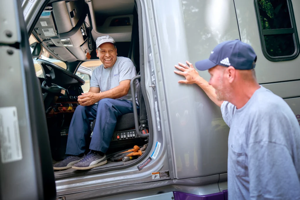 Two men in casual clothes smile and converse; one sits inside a truck cab behind the wheel, while the other stands outside with his hand on the truck door.
