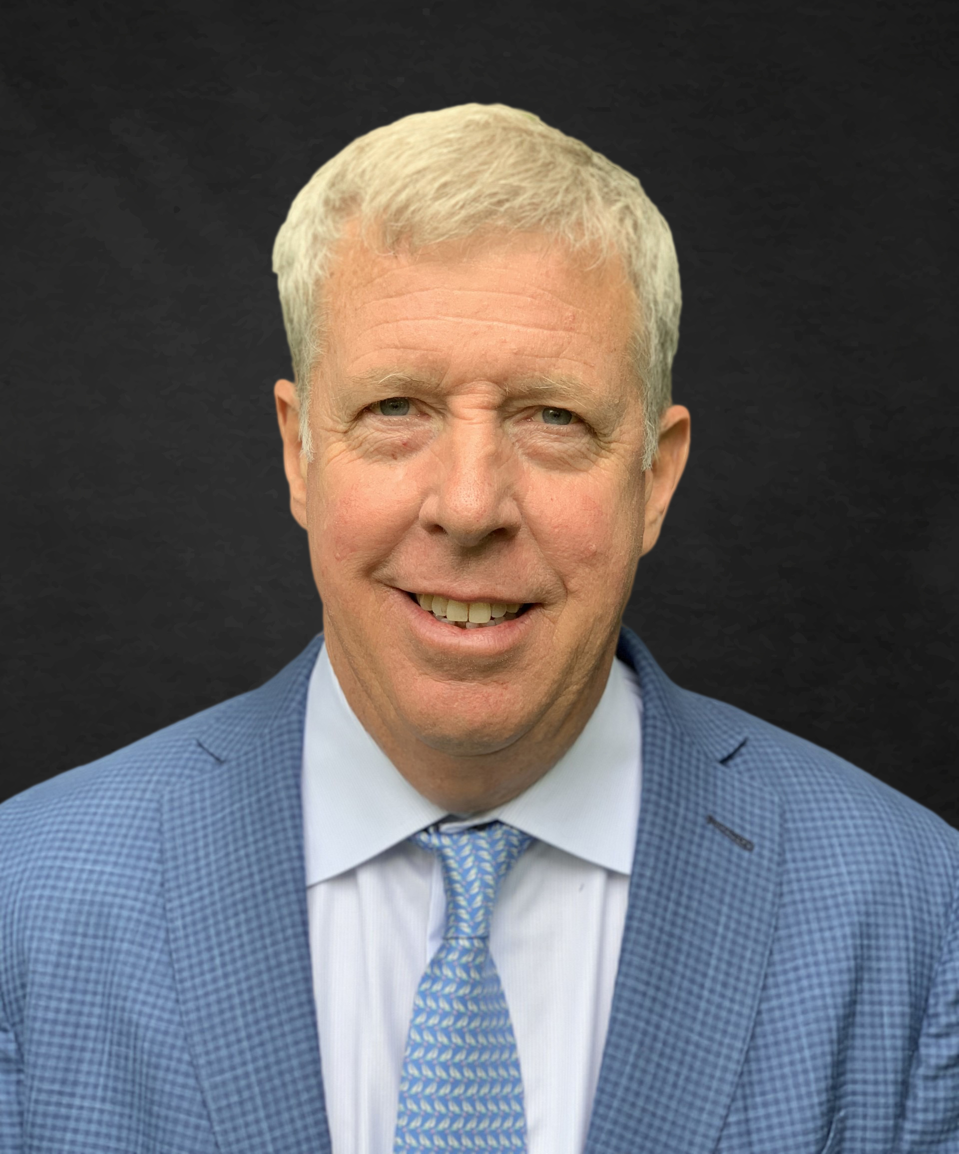 A middle-aged man with short gray hair, wearing a light blue checkered suit jacket, blue dress shirt, and a patterned blue tie, smiles in front of a plain dark background.