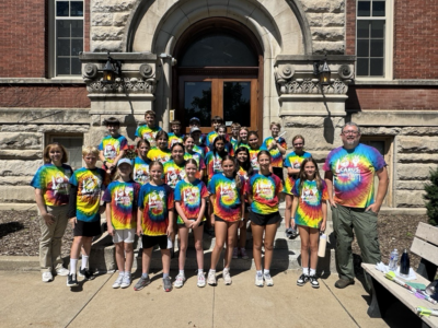 A group of children and two adults wearing colorful tie-dye shirts pose and smile together in front of a historic stone building with arched wooden doors on a sunny day.