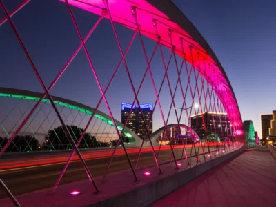 A modern arched bridge in Fort Worth, Texas, is lit with vibrant pink and green lights at dusk. Skyscrapers and city buildings are visible in the background against a deep blue sky.