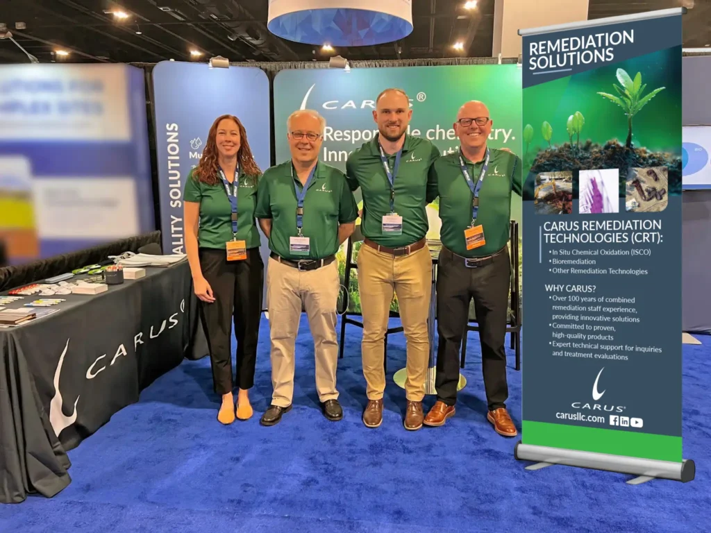 Four people in green polo shirts stand side by side at a trade show booth with a Carus banner about remediation solutions, featuring plant seedlings and informational text. A blue carpet and display tables are visible.