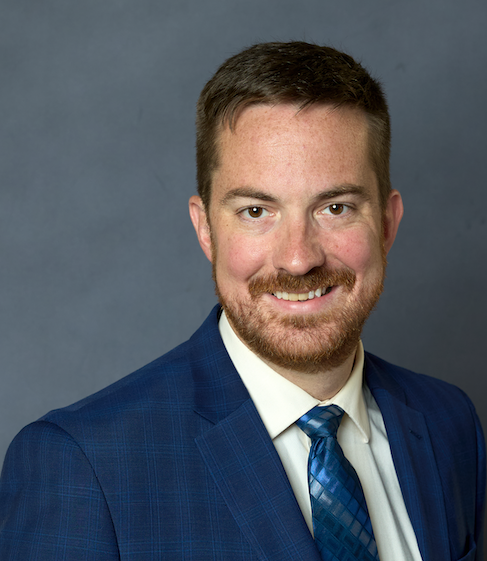 Man with short brown hair and beard, wearing a blue suit, white shirt, and blue patterned tie, smiling in front of a plain gray background.