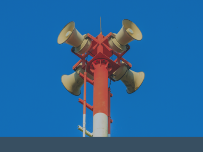 Four yellow loudspeakers are mounted at the top of a red metal pole, facing different directions, against a clear blue sky.