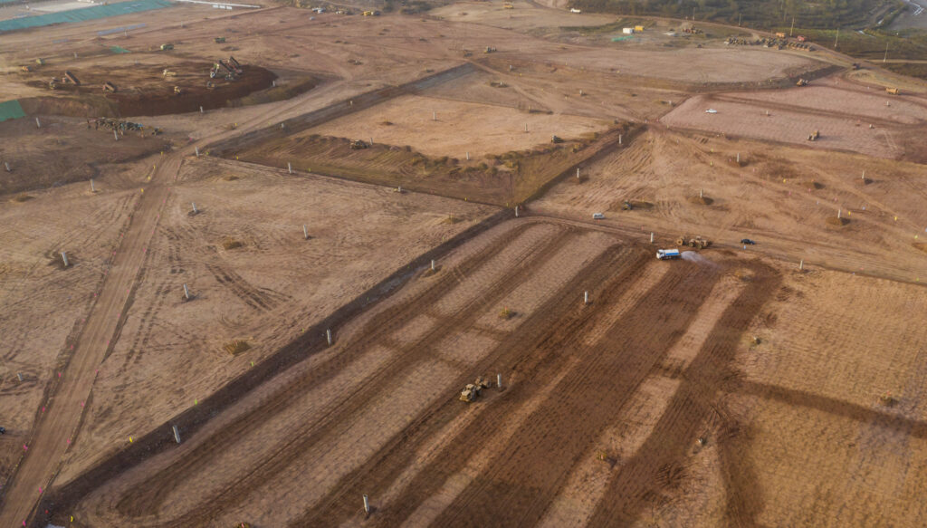 Aerial view of a large construction site with brown dirt fields, machinery, and dirt roads, showing land being cleared and prepared for development.