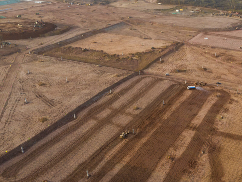 Aerial view of a large construction site with brown dirt fields, machinery, and dirt roads, showing land being cleared and prepared for development.