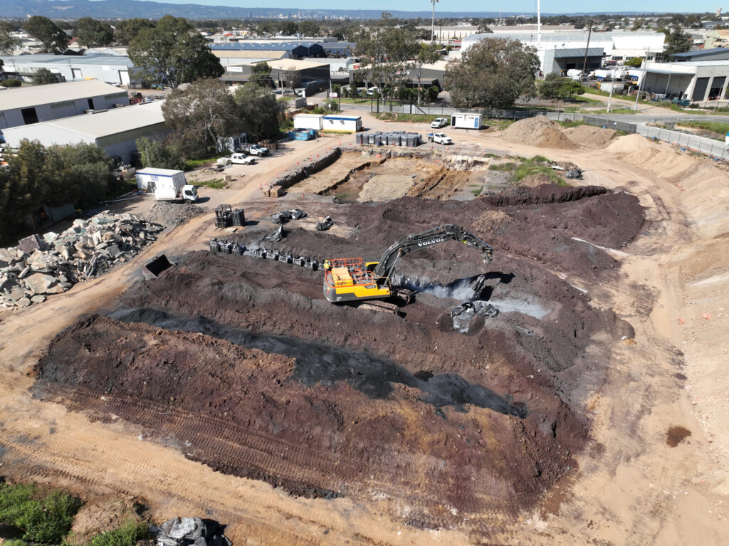 A large yellow excavator digs in a dirt construction site, surrounded by piles of soil, rubble, and some buildings in the background under a clear sky.