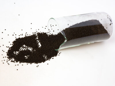 A glass beaker resting on its side with dark brown CARULITE catalyst granules spilling out onto a white surface.