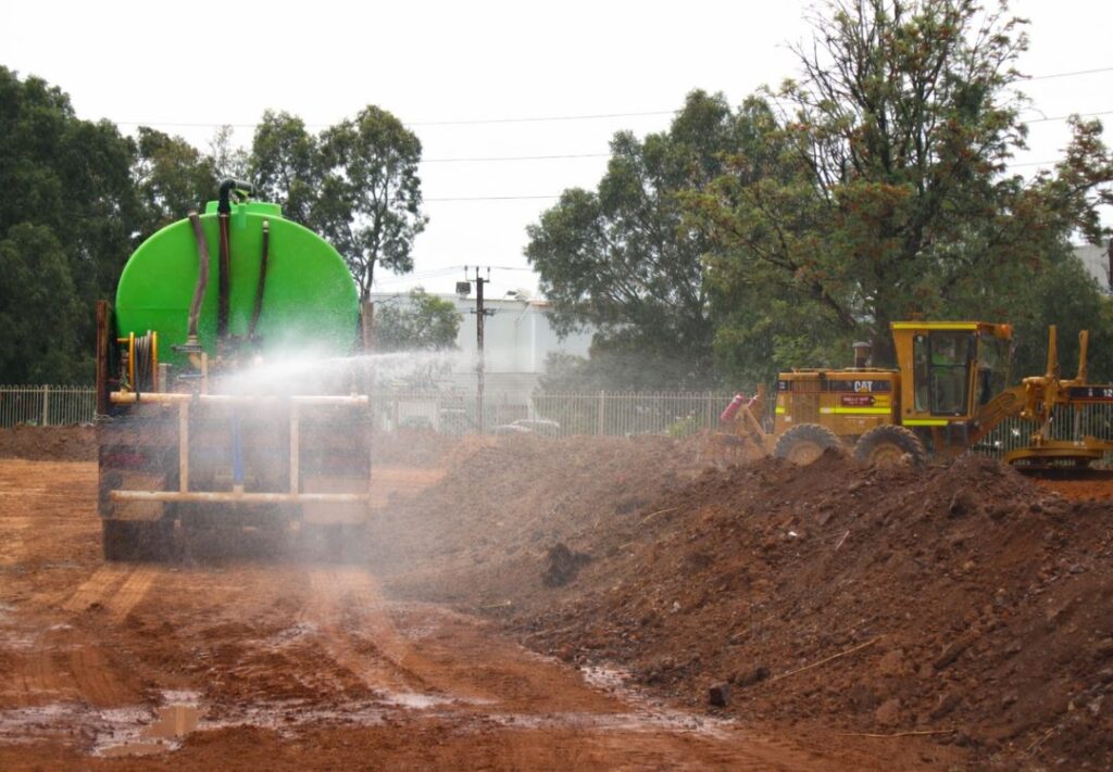A water truck spraying diluted RemActiv over windrowed soil while a grader works the treatment area.