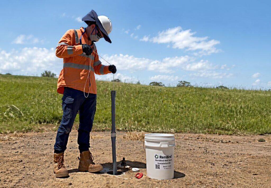 A field technician in hi-vis lowering a RemBind Well Sock chain into a groundwater monitoring well beside a RemBind Well Sock installation kit.